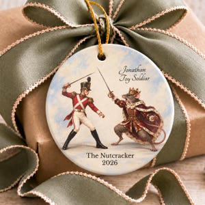 Peut inclure: Un ornement en céramique rond avec une illustration imprimée d'un soldat en uniforme rouge en duel avec un roi souris portant une couronne et une cape. Le texte indique Jonathan Toy Soldiar, The Nutcracker et 2026. Il est suspendu par un cordon doré.