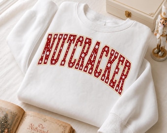 Könnte beinhalten: Ein weißes Langarm-Sweatshirt mit dem Wort NUTCRACKER, das in einer großen, geschwungenen, roten Schrift im College-Stil gedruckt ist. Die Buchstaben sind mit einem wiederkehrenden Muster aus kleinen weißen Schneeflocken gefüllt.