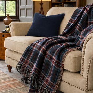 May include: A beige upholstered sofa with nailhead trim features a dark navy blue square throw pillow and a draped woven throw blanket. The blanket displays a plaid pattern in shades of black, deep red, and blue, finished with fringed edges.