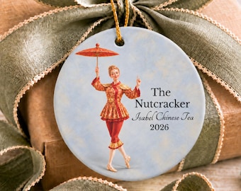 Könnte beinhalten: Ein rundes Keramikornament mit hellblauem Hintergrund zeigt eine tanzende Person in rot-gold gemusterter Kleidung mit einem roten Sonnenschirm. Der Text lautet The Nutcracker, Isabel Chinese Tea und 2026. Es hängt an einer goldenen Schnur.