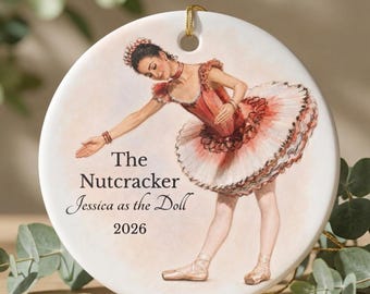 May include: A round white ceramic ornament featuring a watercolor illustration of a ballerina in a pink and white tutu. The text reads The Nutcracker, Jessica as the Doll, and 2026. A gold string is attached to the top for hanging purposes.