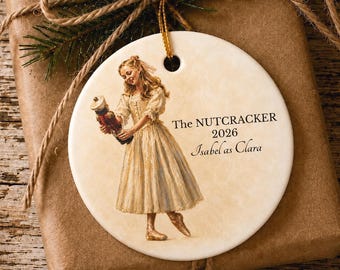 May include: A round white ceramic ornament featuring a printed illustration of a girl in a cream-colored dress holding a nutcracker. The text reads The NUTCRACKER 2026 Isabel as Clara. A gold string is attached to the top for hanging purposes.