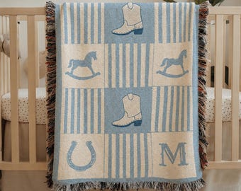 May include: A woven throw blanket featuring a light blue and cream color scheme with a grid pattern. The design includes alternating blocks of vertical stripes and icons of cowboy boots, rocking horses, a horseshoe, and the capital letter M.