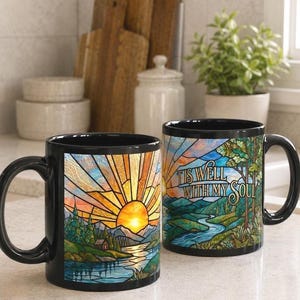 May include: Two black ceramic mugs featuring a stained glass style design of a sunrise over mountains, a river, and a cabin. One mug displays the text 'TIS WELL WITH MY SOUL' in a decorative font, while the other shows the full landscape scene.