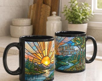 May include: Two black ceramic mugs featuring a stained glass style design of a sunrise over mountains, a river, and a cabin. One mug displays the text 'TIS WELL WITH MY SOUL' in a decorative font, while the other shows the full landscape scene.