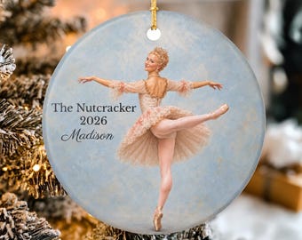 May include: A round ceramic ornament featuring a ballerina in a light pink tutu performing a dance pose. The text "The Nutcracker 2026 Madison" is printed on the left side. The ornament has a gold string loop at the top for hanging purposes.