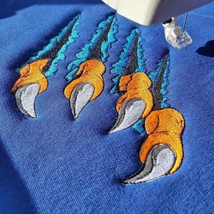 Puede incluir: Primer plano de una tela azul con un diseño bordado de garras naranjas y grises rasgando un fondo azul y turquesa. La máquina de coser es blanca.