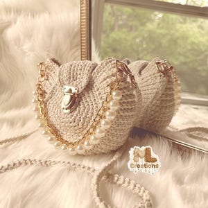 May include: A beige crochet handbag with a gold chain and pearl accents. The bag has a gold clasp and a matching crochet strap. The bag is reflected in a mirror. The bag has the text "ML creations".