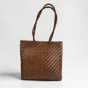 Può includere: Una borsa tote intrecciata marrone con manici intrecciati. La borsa ha una forma quadrata e un motivo a trama diagonale strutturato. I manici sono anch'essi marroni e intrecciati, su sfondo bianco.