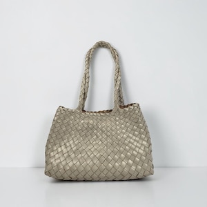 Può includere: Una borsa tote intrecciata di colore grigio chiaro neutro. La borsa ha una forma rettangolare con un manico intrecciato. Il motivo intrecciato è uniforme su tutto il corpo della borsa, creando una superficie strutturata. La borsa è adatta all'uso quotidiano.