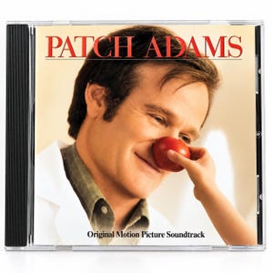 May include: A CD case for the Patch Adams Original Motion Picture Soundtrack. The cover features a man in a white coat and olive green shirt with a red tomato on his nose, held in place by a hand. The text reads PATCH ADAMS at the top.