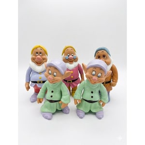 Puede incluir: Un conjunto de cinco figuras de plástico que representan personajes con orejas puntiagudas, vistiendo túnicas, cinturones y gorros de varios colores, incluyendo verde claro, morado, rosa, azul y marrón, cada una con expresiones faciales únicas.