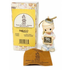 May include: A porcelain ornament featuring a small figure with blonde hair, a silver halo, and a white robe, holding a small chalkboard with the number 96. The item includes a yellow cardboard box labeled Peace On Earth...Anyway 1996 Annual Edition.