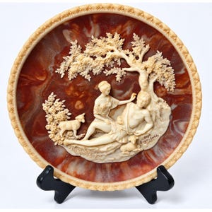 May include: A round decorative plate with a deep reddish-brown marbled center and a cream-colored border. The center features a raised cream-colored relief of two figures resting under a tree with two small animals. The text Gayle Appleby is visible.