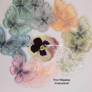 May include: Embroidered butterfly appliques in pastel shades of blue, pink, orange, and gold. Each butterfly has detailed wing patterns and a central body. A pansy flower is also present.