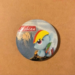 May include: A round button with a light blue pony figure with a rainbow mane and tail, set against a mountain landscape. The word "Lesbian" is printed in red on the button.