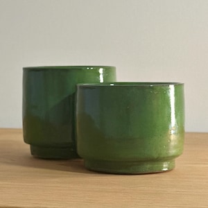 May include: Two green ceramic cups with a glossy finish. The cups are cylindrical with a slightly flared base, and are set on a wooden surface. The cups are a vibrant shade of green.