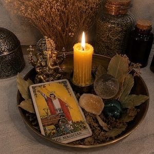 May include: A brass tray containing a lit yellow pillar candle, a gold-toned Ganesha figurine, a The Magician tarot card, a clear crystal sphere, a rough yellow stone, a dark green polished stone, and dried bay leaves arranged in a cluster.