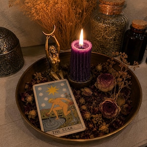 May include: A round brass tray containing a purple pillar candle with a lit flame, a gold-toned figurine of a person holding a crescent moon, dried rosebuds, and a tarot card labelled The Star with the Roman numeral XVII at the top.