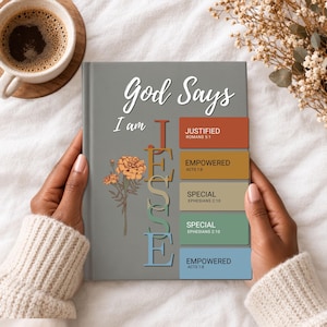 May include: A gray hardcover journal featuring the text God Says I am JESSE in white and colorful lettering. The design includes a marigold flower illustration and five rectangular blocks with the words JUSTIFIED, EMPOWERED, and SPECIAL.