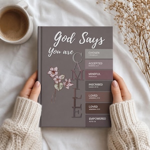 May include: A hardback book with a dusty purple cover featuring the white text God Says You are, the name CAMILLE written vertically, and a floral illustration. To the right, a list of words includes CHOSEN, ACCEPTED, MINDFUL, INSCRIBED, LOVED, and EMPOWERED.