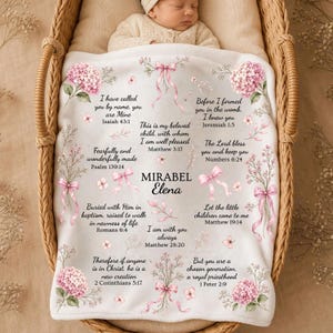 Peut inclure: Une couverture pour bébé blanche ornée d'illustrations d'hortensias roses, de rubans rose clair et de divers versets bibliques imprimés en noir. Le centre de la couverture affiche le nom Mirabel Elena dans une police d'écriture décorative.