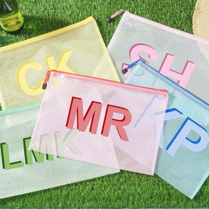 Peut inclure: Cinq pochettes zippées en maille de couleurs pastel : jaune, rose, bleu, vert et rose. Chaque pochette porte un ensemble différent de grandes lettres imprimées. Les pochettes sont disposées sur de l'herbe verte.