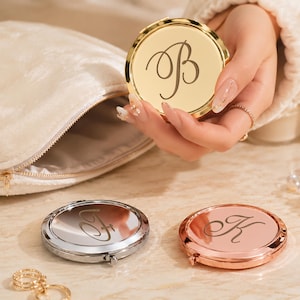 Pode incluir: Um espelho compacto dourado com a letra "B" em fonte cursiva é segurado na mão. Dois outros espelhos compactos, um prateado e outro ouro rosa, estão em uma superfície. Uma bolsa de cosméticos creme e maquiagem também são visíveis.