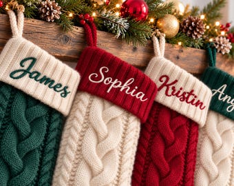 Op de afbeelding: Een rij van vier gebreide kerstsokken met kabelpatronen en geribbelde boorden. De sokken zijn gepersonaliseerd met geborduurde namen: James in donkergroen, Sophia in wit, Kristin in rood en Ange in wit.