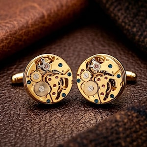 May include: Gold-coloured cufflinks showcasing the inner workings of a watch. The round cufflinks have intricate gears, blue detailing, and a polished finish. They are presented on a brown leather surface.