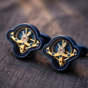 May include: A pair of blue cufflinks with a watch mechanism design. The cufflinks feature a dark blue frame with gold-colored gears and a clear center, showcasing the intricate watch movement. The background is a textured gray fabric.