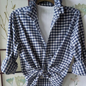 May include: A long-sleeved button-down shirt featuring a navy blue and white gingham check pattern. The shirt has a ruffled collar, a front button closure, and is tied at the waist. It is layered over a plain white undershirt with rolled-up sleeves.