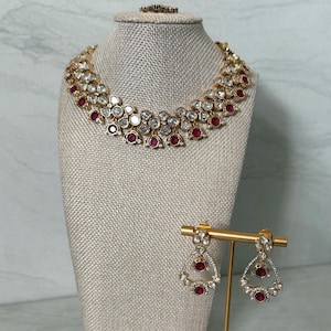 May include: A gold-toned necklace featuring multiple rows of clear, faceted stones interspersed with round, deep red stones. The set includes matching drop earrings, each with a central red stone surrounded by clear stones, displayed on a gold stand.