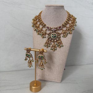 May include: A gold-toned necklace and matching earring set featuring intricate metalwork, circular green enamel centers, small pink stones, and dangling white bead accents. The necklace is displayed on a fabric bust, while the earrings hang from a gold stand.