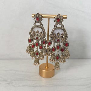 May include: A pair of ornate chandelier earrings featuring a gold-tone metal frame, white enamel accents, and multiple oval-shaped red stones. The design includes intricate floral detailing and small dangling bell-shaped charms at the base.