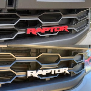 May include: A black automotive front grille featuring a prominent, raised emblem with the text RAPTOR. The image displays two variations of the emblem, one in a bold red finish and the other in a clean white finish, both mounted on the honeycomb structure.