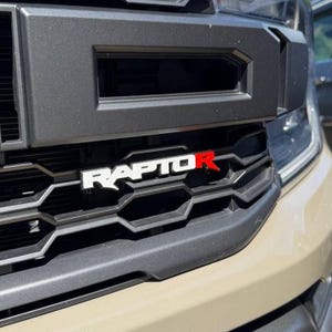 May include: A matte black vehicle front grille featuring a prominent honeycomb pattern and a bold emblem that reads RAPTOR with the letter R highlighted in red. The grille is integrated into a tan-colored vehicle body with a smooth finish.