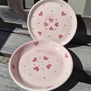 Peut inclure: Deux petites assiettes rondes en céramique rose. Chaque assiette présente un motif de cœurs et de points rose foncé. Les assiettes sont posées sur une surface en bois, et l'éclairage est vif.