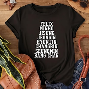 May include: Black t-shirt with white text listing the names: Felix, Minho, Jisung, Jeongin, Hyunjin, Changbin, Seungmin, and Bang Chan. The shirt is displayed on a wooden surface with a cowboy boot, hat, and sunglasses.