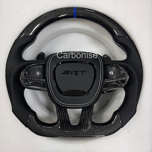 Puede incluir: Un volante negro y de fibra de carbono con una franja azul en la parte superior. El centro presenta el texto "SRT" y "Carbonise" en blanco. El volante tiene empuñaduras de cuero negro perforado y costuras blancas.