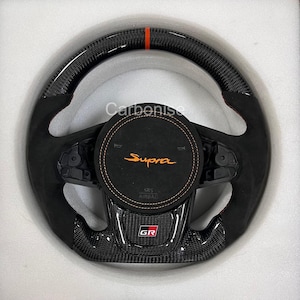 May include: A black and carbon fibre steering wheel with orange stitching and accents. The centre features the word "Supra" in orange, with the "GR" logo below. The wheel has a sporty design.