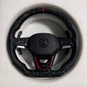 Puede incluir: Un volante negro y de fibra de carbono con una raya roja. El volante presenta un centro negro con el logotipo de Volkswagen y controles integrados. El borde tiene un agarre de cuero perforado.