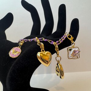 Puede incluir: Una pulsera de dijes dorada con una cadena morada. Los dijes incluyen un coraz&oacute;n, una mariposa, un cuadrado con una flor y un dije redondo. La pulsera se muestra en una mano negra.