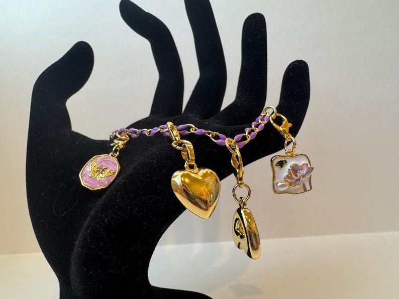Puede incluir: Una pulsera de dijes dorada con una cadena morada. Los dijes incluyen un coraz&oacute;n, una mariposa, un cuadrado con una flor y un dije redondo. La pulsera se muestra en una mano negra.