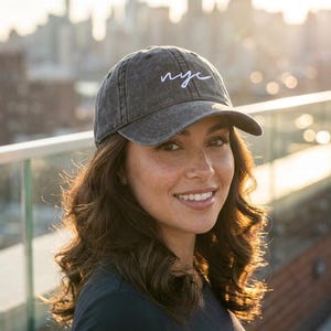 May include: A charcoal grey baseball cap with a distressed, washed-out finish. The front of the cap features the letters nyc embroidered in a white, cursive script font. The cap has a curved brim and a structured crown with a button on top.