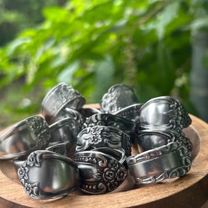May include: A collection of silver-toned metal rings crafted from repurposed spoon handles, featuring various embossed floral and decorative patterns. The rings are arranged in a circular wooden bowl, showcasing intricate details and polished surfaces.