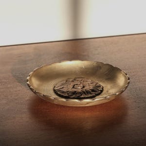May include: A gold-colored, scalloped-edge dish with a detailed, dark brown floral centerpiece. The dish sits on a dark brown wooden surface, with soft lighting creating shadows.