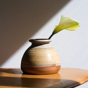 May include: A small, handcrafted ceramic vase with a light brown and dark brown color scheme. A single white and yellow calla lily flower is placed in the vase. The vase sits on a wooden table, with sunlight casting shadows.
