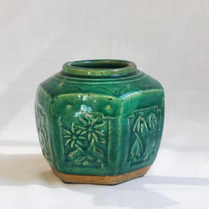 May include: A hexagonal ceramic jar with a glossy emerald green glaze and a visible crackle finish. The exterior features embossed floral panels on each side, with a wide circular opening at the top and a plain, unglazed tan base.
