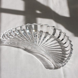May include: A clear glass dish shaped like a fan or a seashell, featuring a series of radiating vertical ridges that extend from the centre to the scalloped outer edge. The glass is transparent and reflects light, creating a bright, faceted appearance.